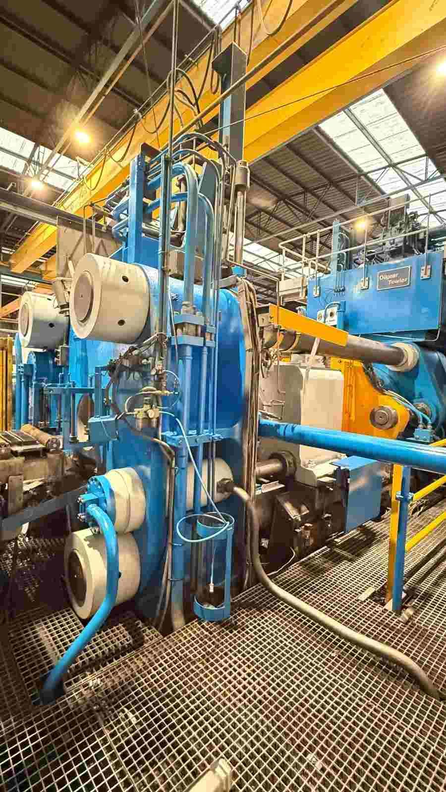 Maquinaria industrial azul en el interior de una fábrica, con una grúa puente amarilla y un suelo de rejilla metálica