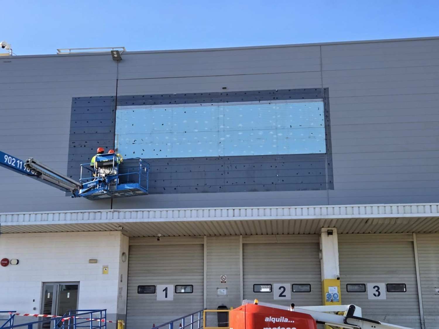 Trabajador subido a una plataforma elevadora pintando un gran panel oscuro en la pared sobre las puertas del muelle de carga