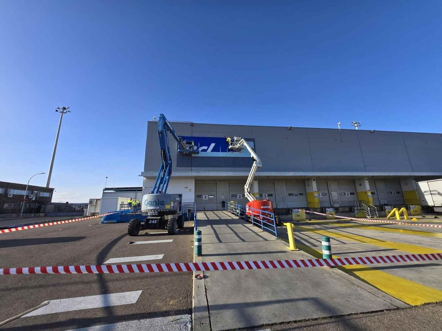 Edificio industrial con una plataforma elevadora de brazo azul y camiones estacionados afuera bajo un cielo despejado.