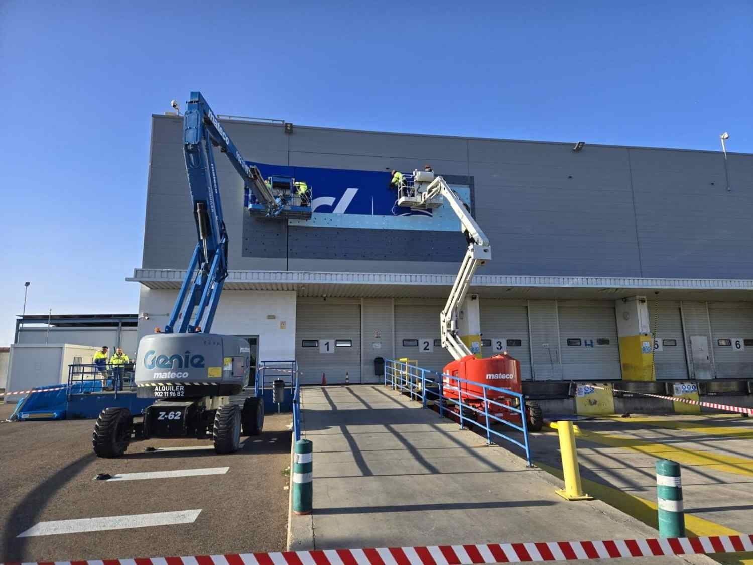Dos trabajadores en una plataforma elevadora limpiando la fachada de un gran almacén bajo un cielo azul.