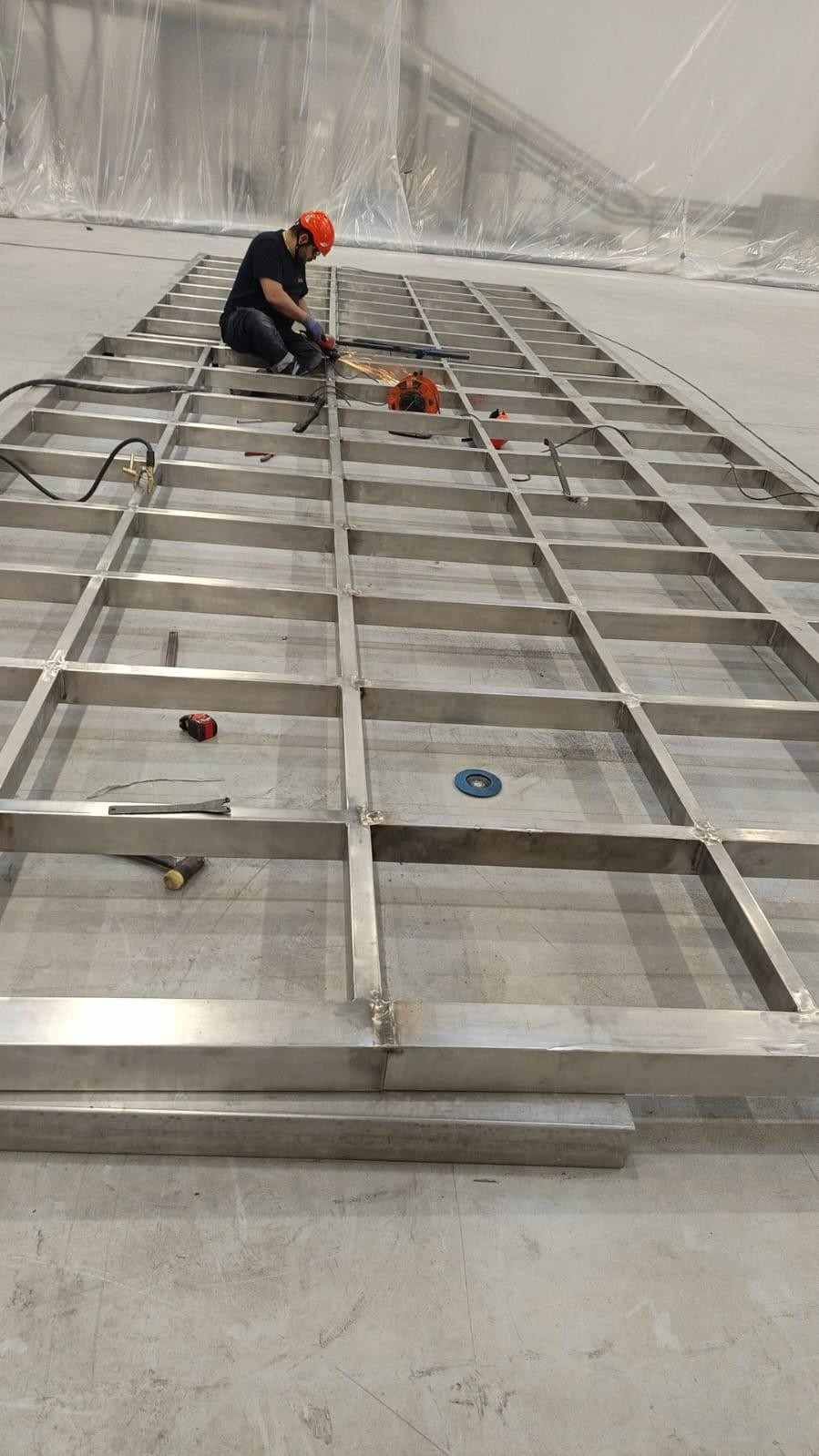 Un trabajador con casco naranja montando una estructura metálica en un amplio espacio industrial.