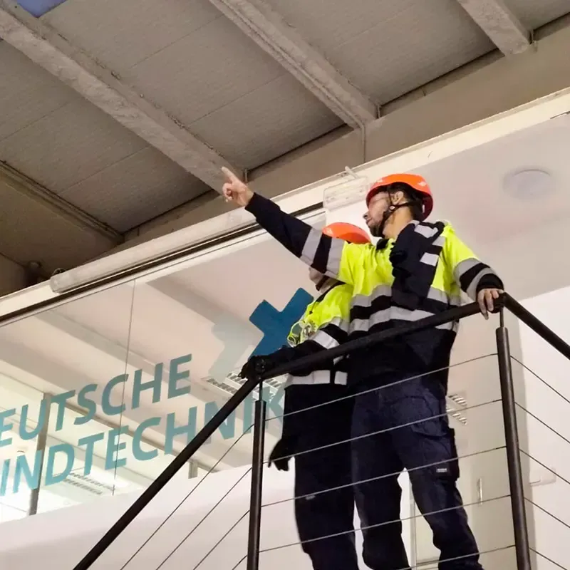Dos obreros de la construcción en una escalera metálica. Uno apunta hacia arriba; ambos llevan equipo de seguridad y chalecos reflectantes.