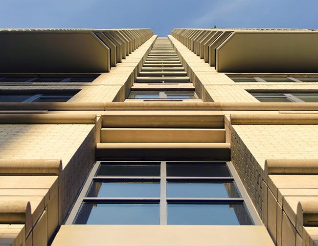 Je lève les yeux vers un grand immeuble en briques beiges avec des balcons et un ciel bleu au-dessus.