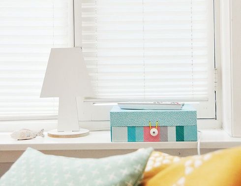 Une lampe blanche, une boîte décorée et une télécommande sont posées sur le rebord d'une fenêtre où l'on aperçoit des stores, un coquillage et des coussins.