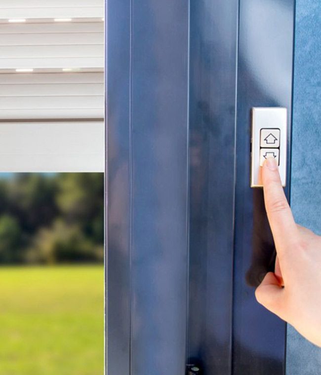 Une personne appuie sur le bouton d'abaissement d'un interrupteur argenté pour baisser un store devant un champ verdoyant.