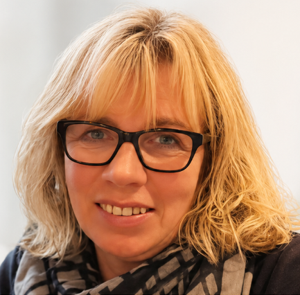 Eine Frau mit Brille und Kopftuch lächelt vor einem Fenster.