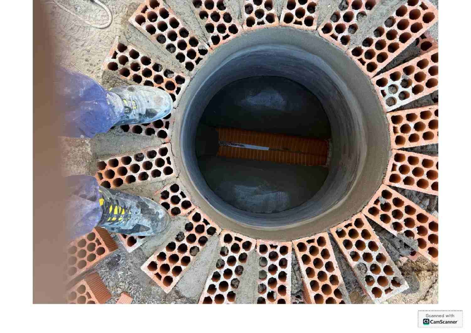 Primer plano de un tubo de desagüe circular dentro de una rejilla metálica perforada, con dos zapatos cerca de la abertura.