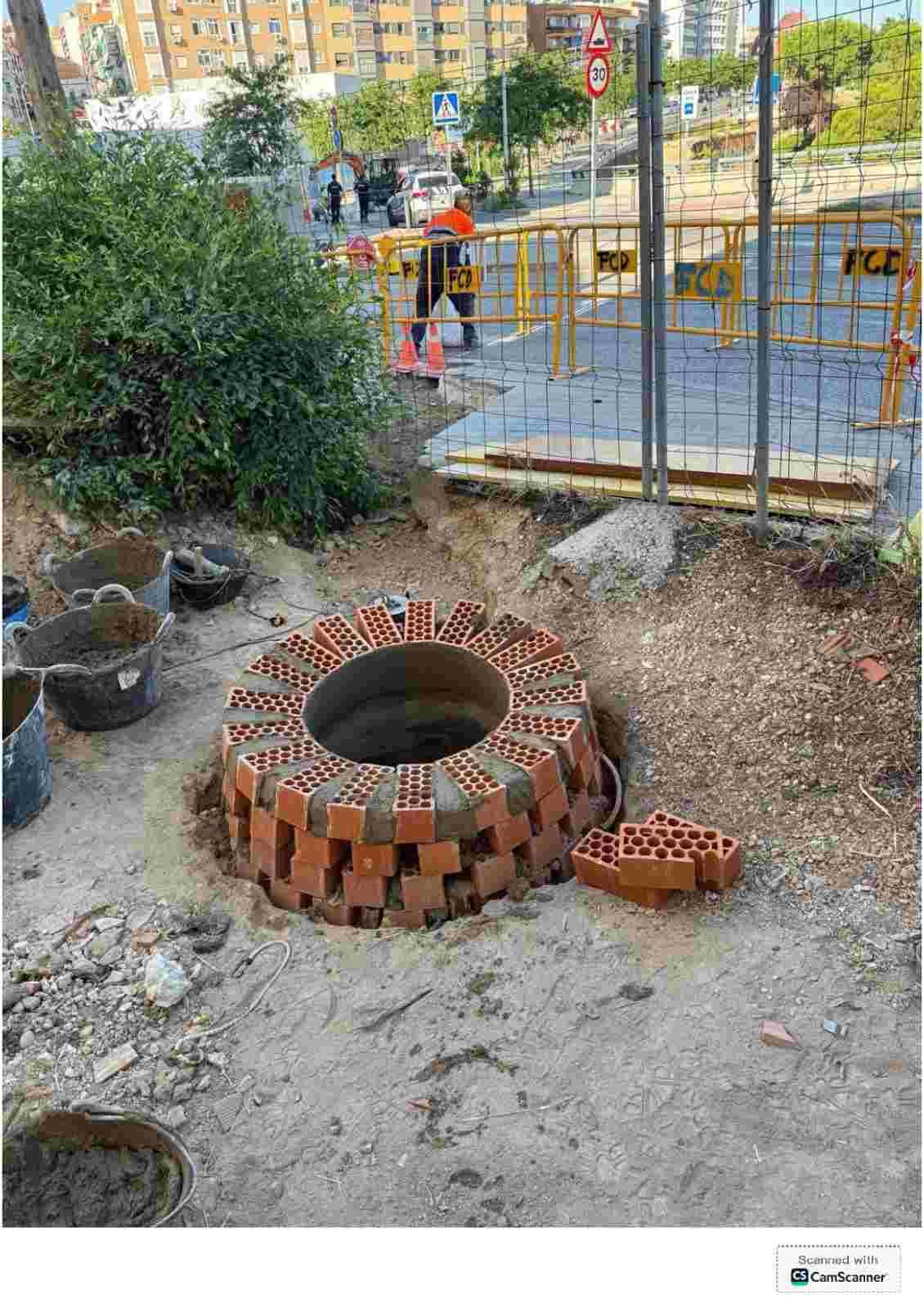 Pozo de ladrillo en construcción en un terreno de tierra, con varillas de acero y trabajadores al fondo.
