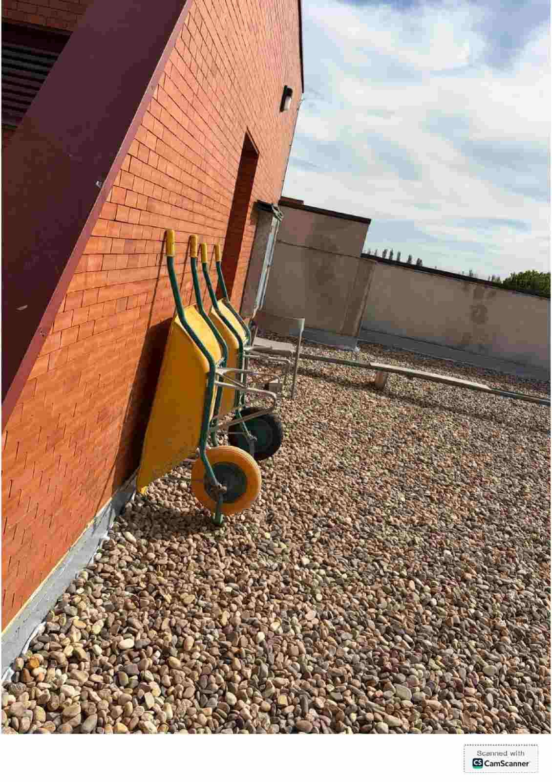 Carretilla de mano amarilla apoyada contra una pared de ladrillo rojo sobre una superficie de grava al aire libre