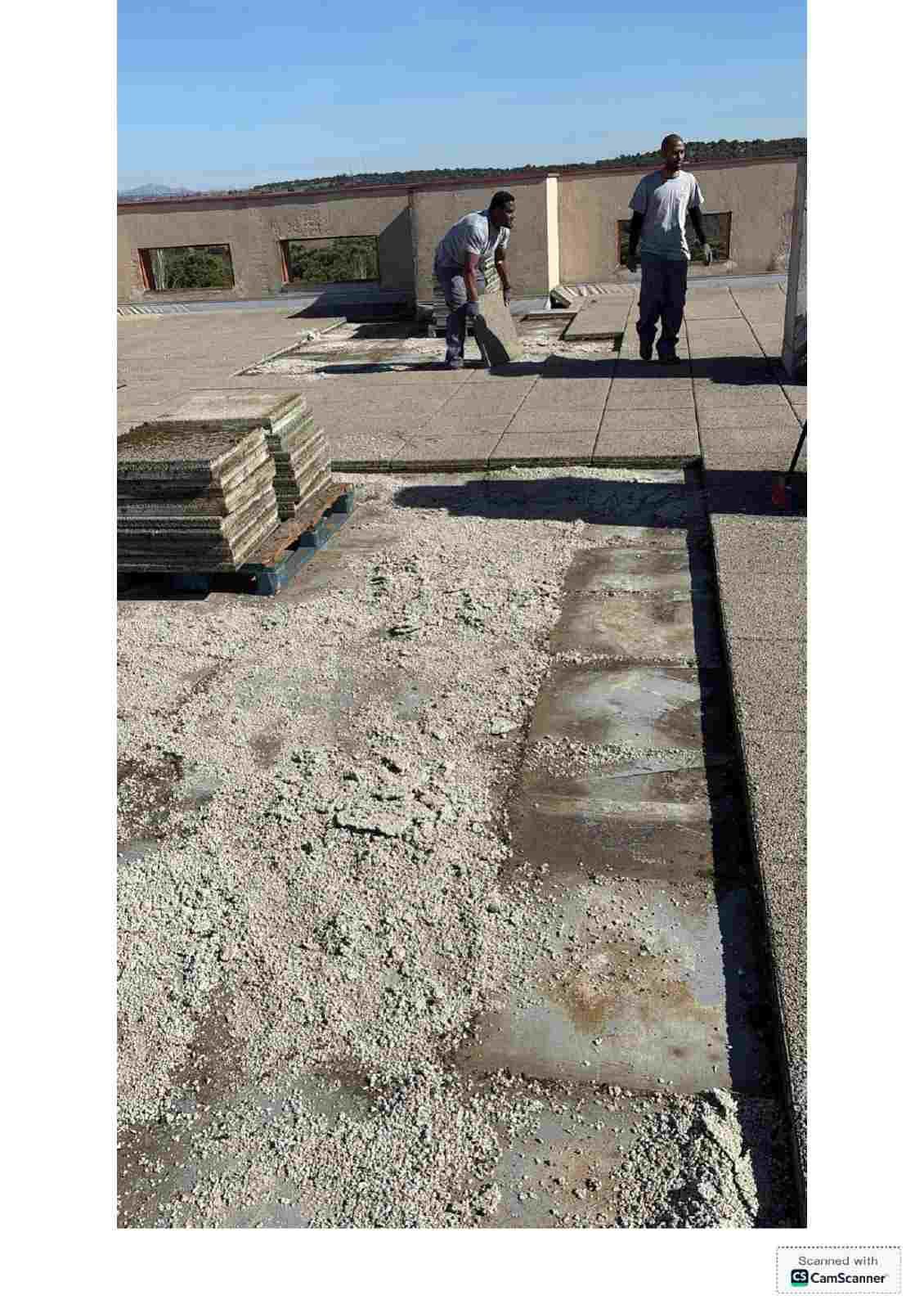 Dos trabajadores en un tejado de grava junto a materiales apilados, bajo un cielo despejado
