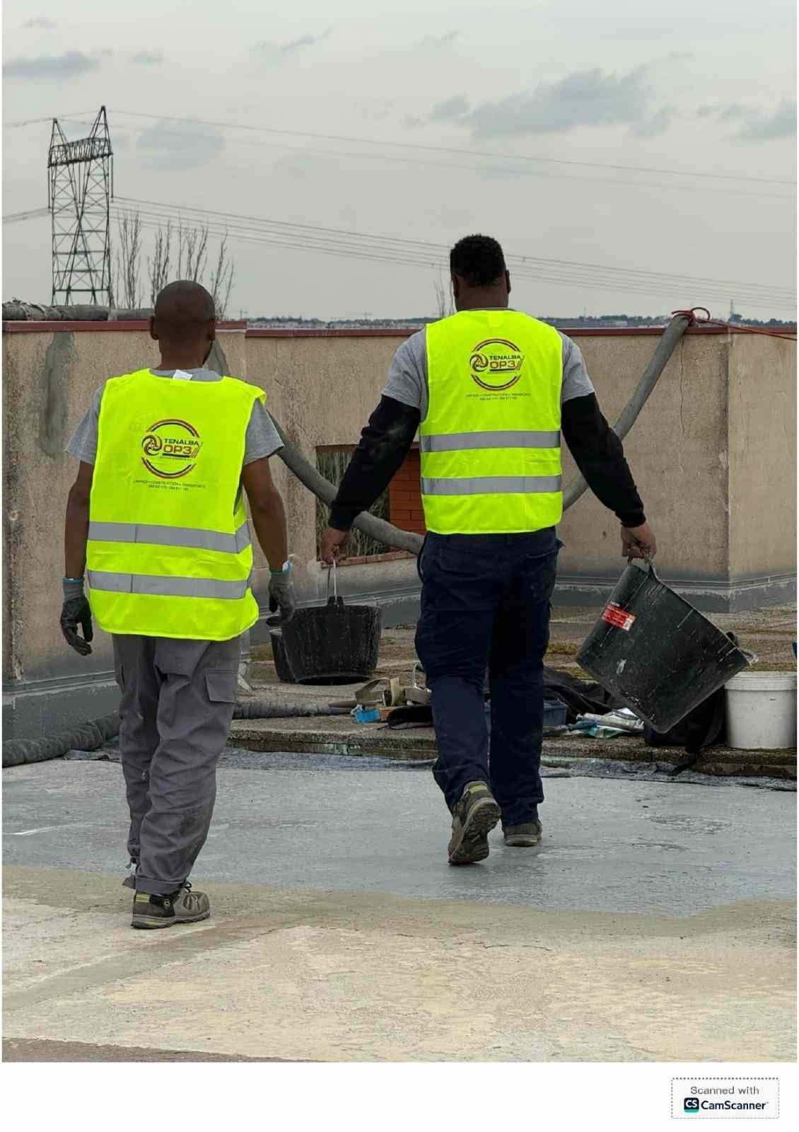 Dos trabajadores con chalecos de seguridad fluorescentes llevan cubos por una obra mojada.