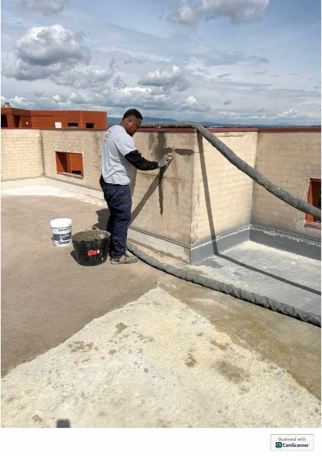 Un trabajador aplicando sellador a un peto de azotea junto a cubos y herramientas, bajo un cielo nublado