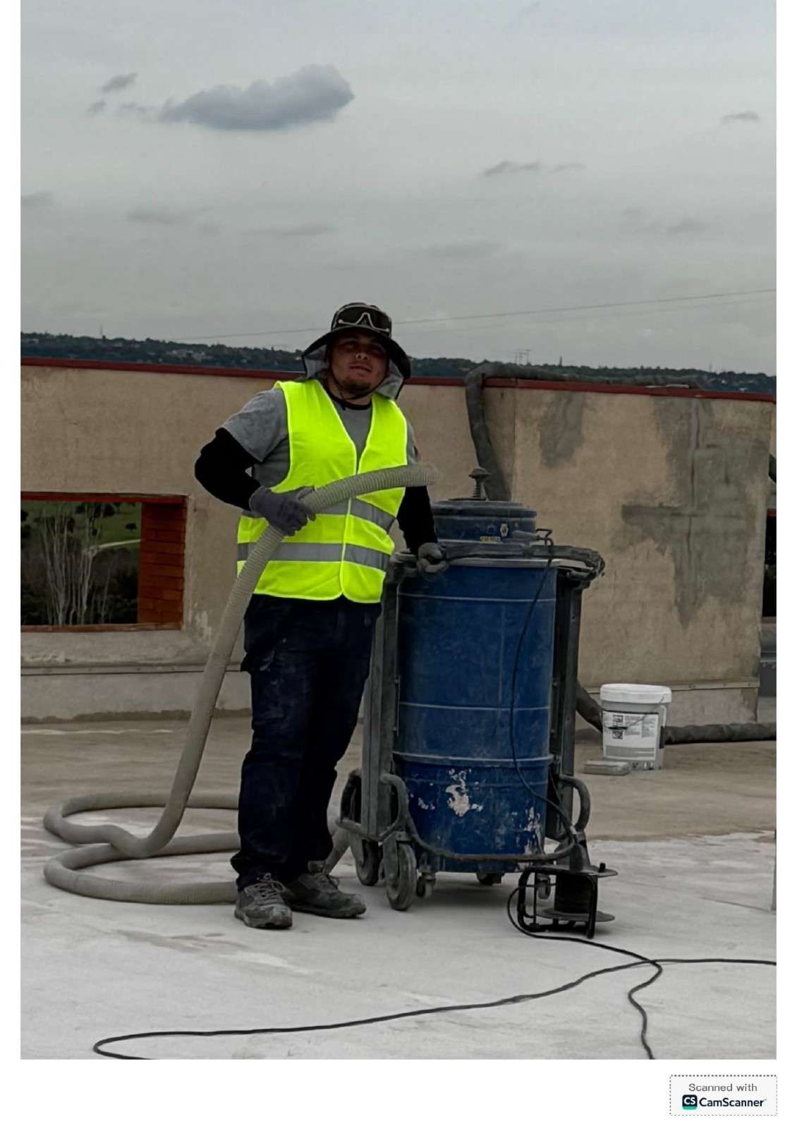 Un trabajador con chaleco reflectante está de pie junto a una aspiradora industrial azul en la azotea de una obra.