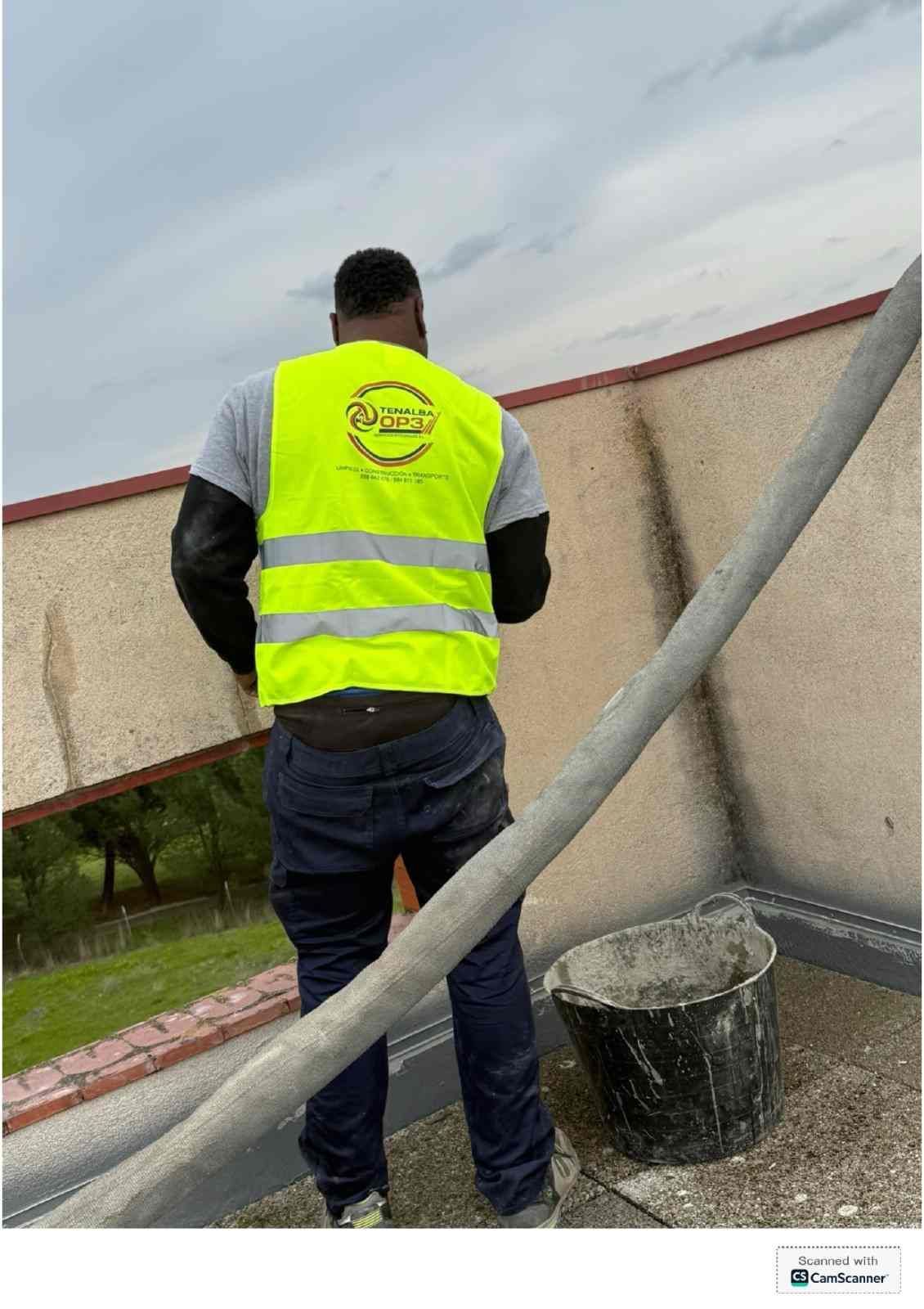 Trabajador con un chaleco amarillo fluorescente en un tejado, junto a una pared y una tubería de ventilación.
