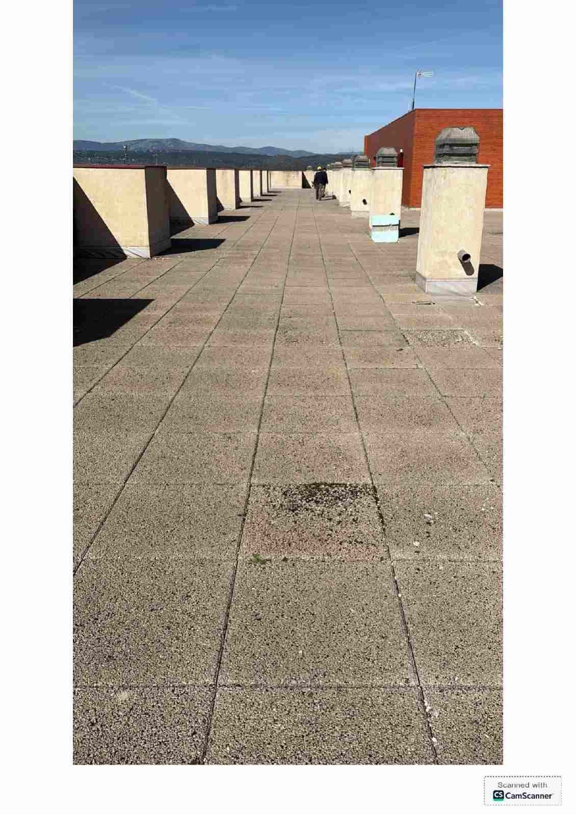 Pasarela en la azotea con suelo de baldosas, pilares de hormigón y un edificio rojo bajo un cielo azul despejado.