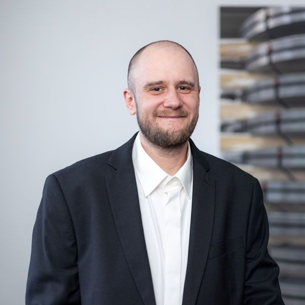 Ein Mann mit Bart trägt ein kariertes Hemd und lächelt in die Kamera.