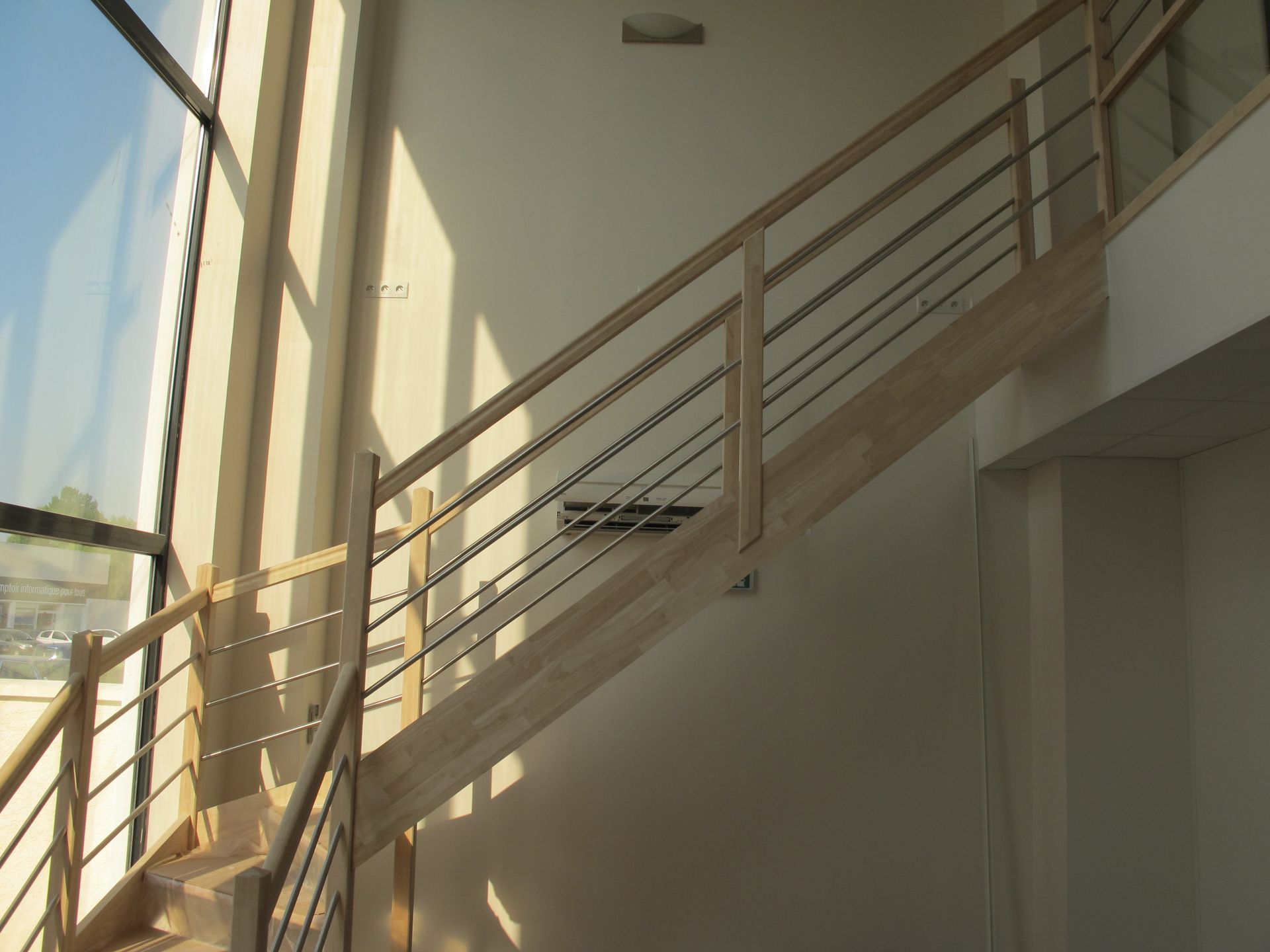 Escalier intérieur baigné de lumière naturelle, avec rampes beiges et grandes fenêtres le long du mur de gauche.