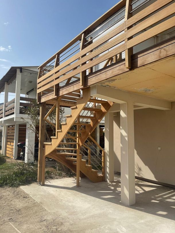 Escalier extérieur en bois et terrasse sous un bâtiment beige, avec des colonnes de soutien blanches et un ciel bleu.