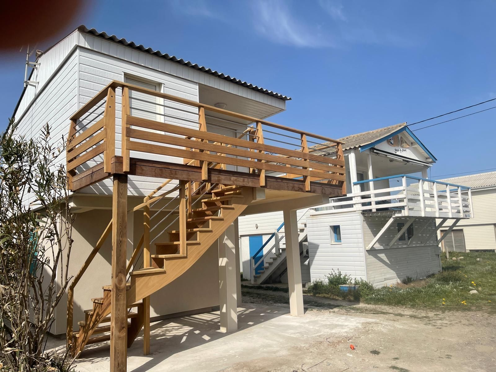 Terrasse en bois surélevée et escalier attenant à une maison blanche sous un ciel bleu azur.