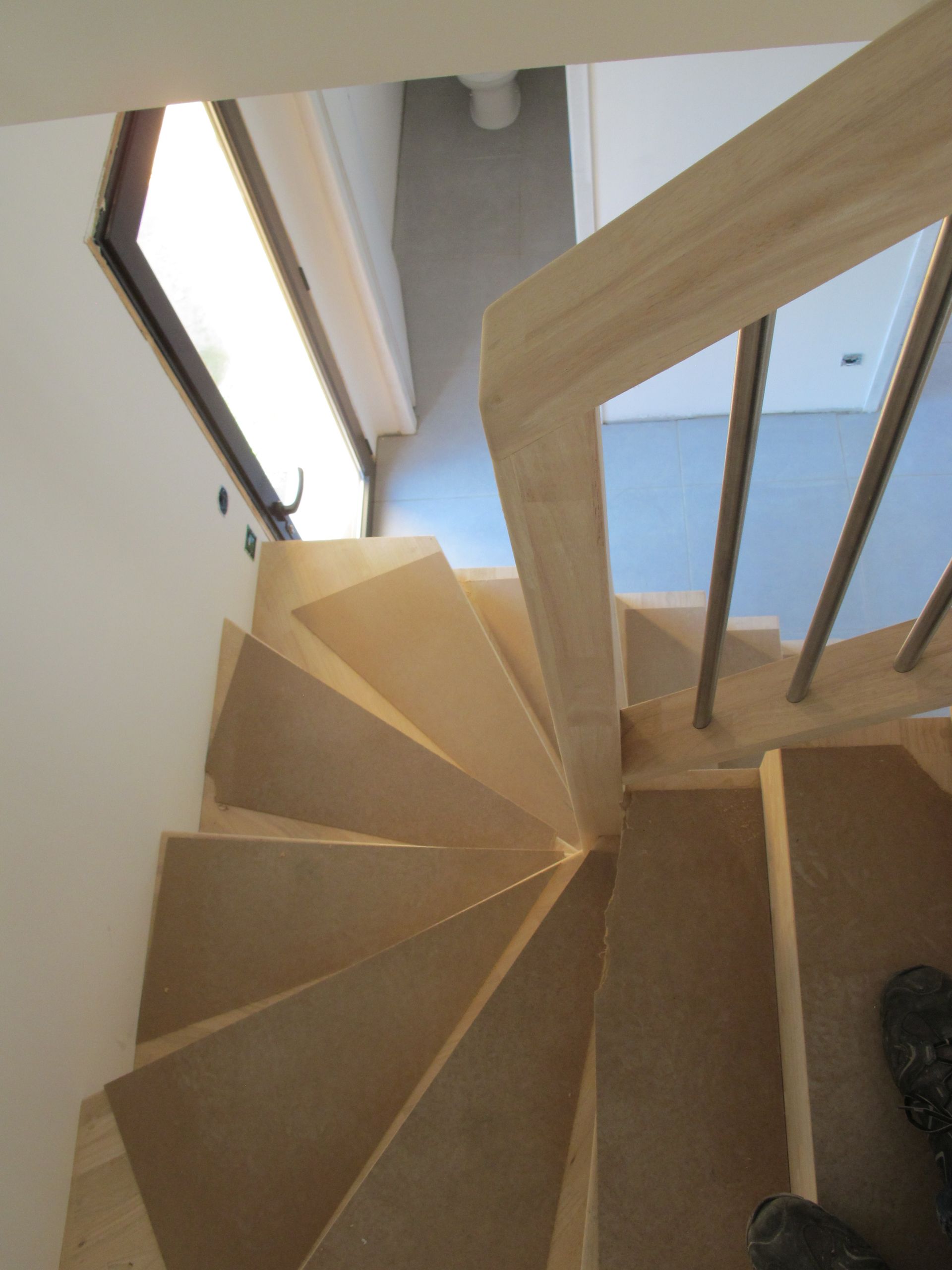Vue plongeante d'un escalier intérieur étroit aux marches en bois clair et aux murs blancs.