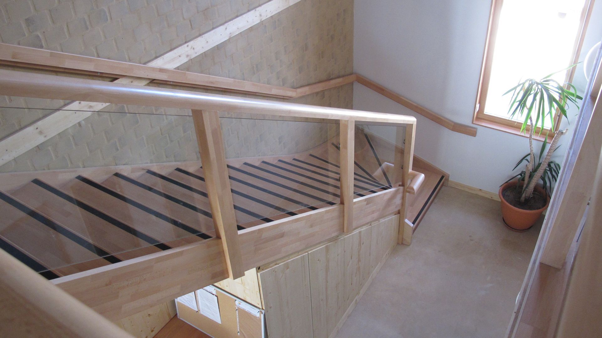 Palier d'escalier en bois avec rampe dans un couloir intérieur lumineux, à côté d'une plante en pot près de la fenêtre