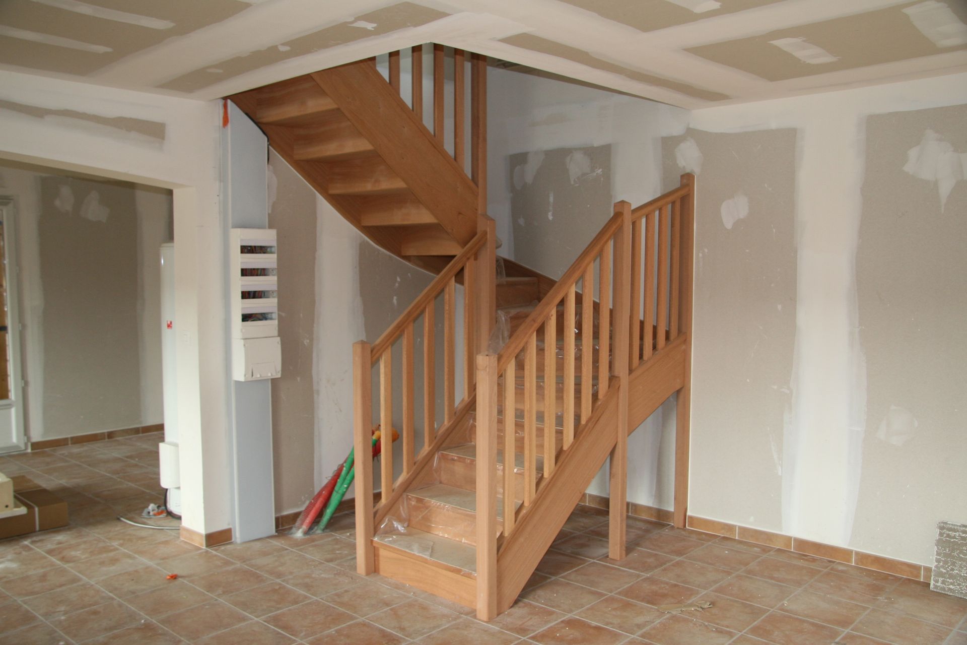 Escalier en bois dans une pièce inachevée avec des plaques de plâtre réparées et un sol carrelé.