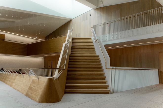 Un escalier en bois avec une rampe est adossé à un mur blanc sur un sol carrelé, avec un balai à proximité.