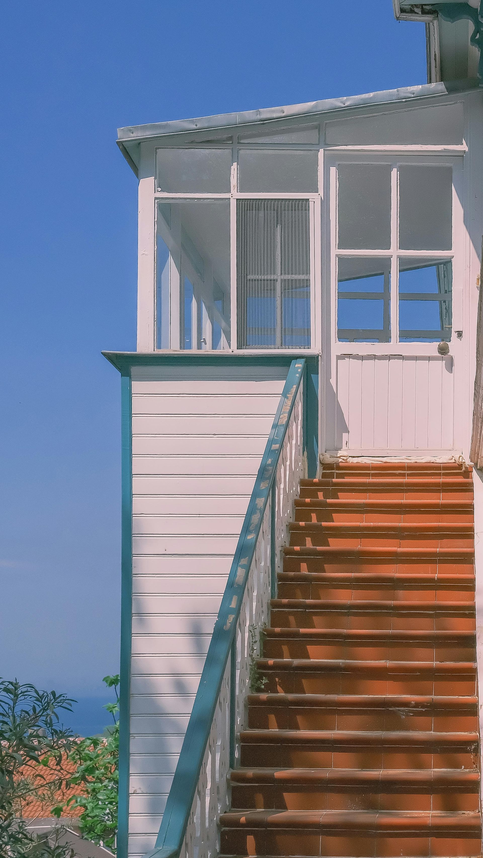 Un escalier en bois mène au porche d'une maison blanche aux fenêtres grillagées, sous un ciel bleu.