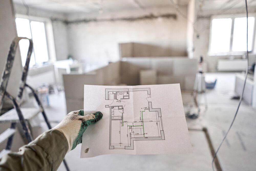 Mano sosteniendo un plano de una habitación en construcción; fondo de un interior polvoriento e inacabado con una escalera.