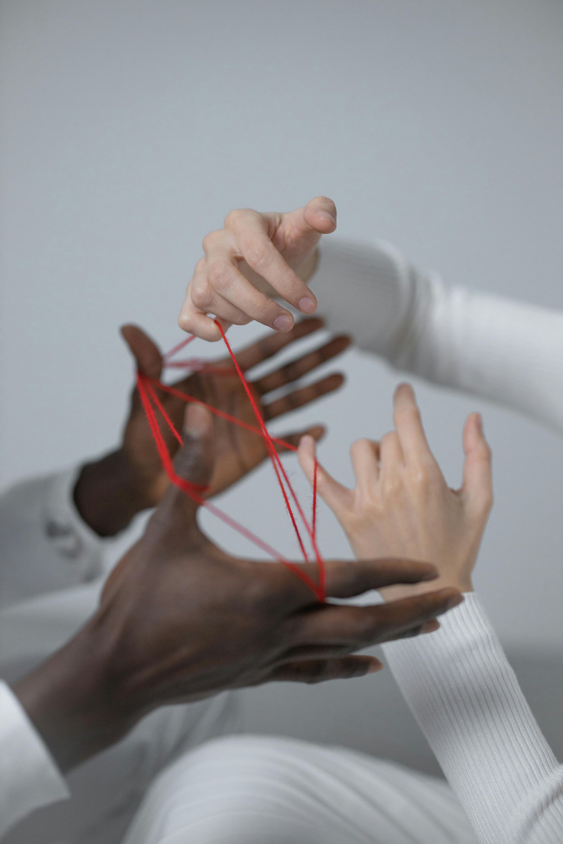 Die Hände dreier Personen mit unterschiedlichen Hauttönen spielen Fadenspiele mit roter Schnur.