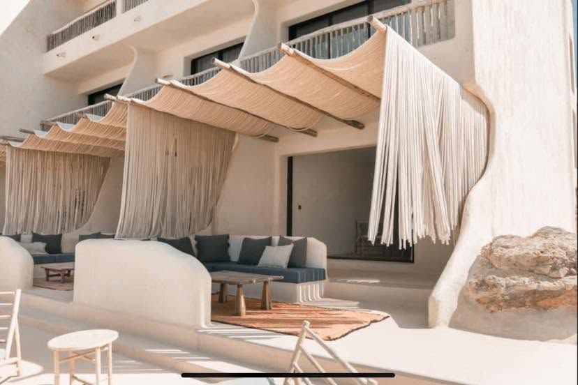 Salón exterior sombreado con toldo beige, asientos blancos y un muro de piedra en un patio soleado.