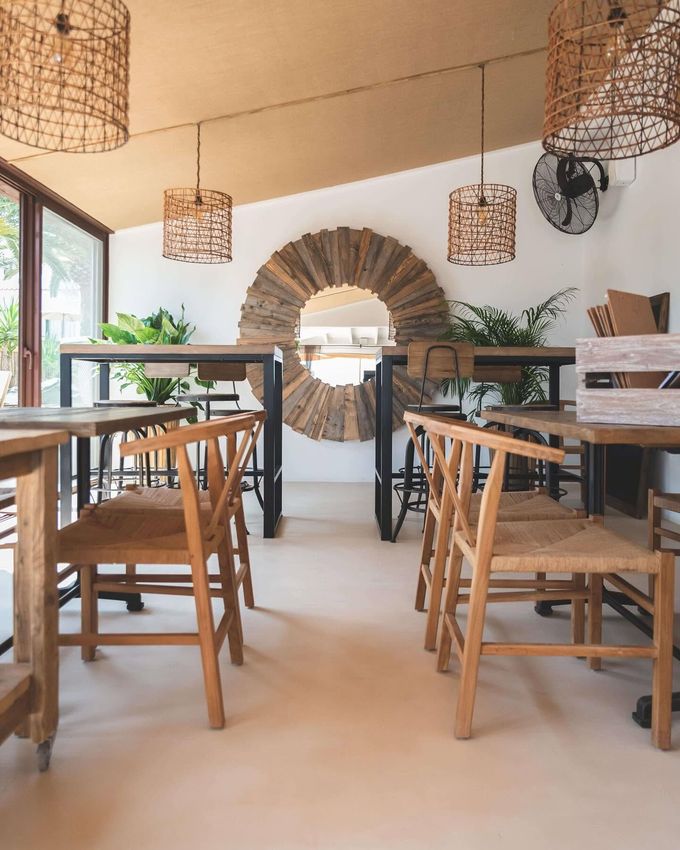 Interior luminoso de la cafetería con mesas y sillas de madera, lámparas colgantes tejidas y un espejo redondo en la pared.