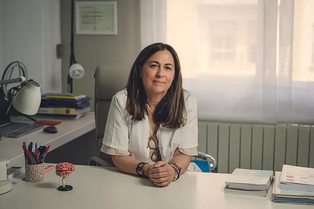 Una mujer está sentada en un escritorio con las manos juntas y sonriendo.