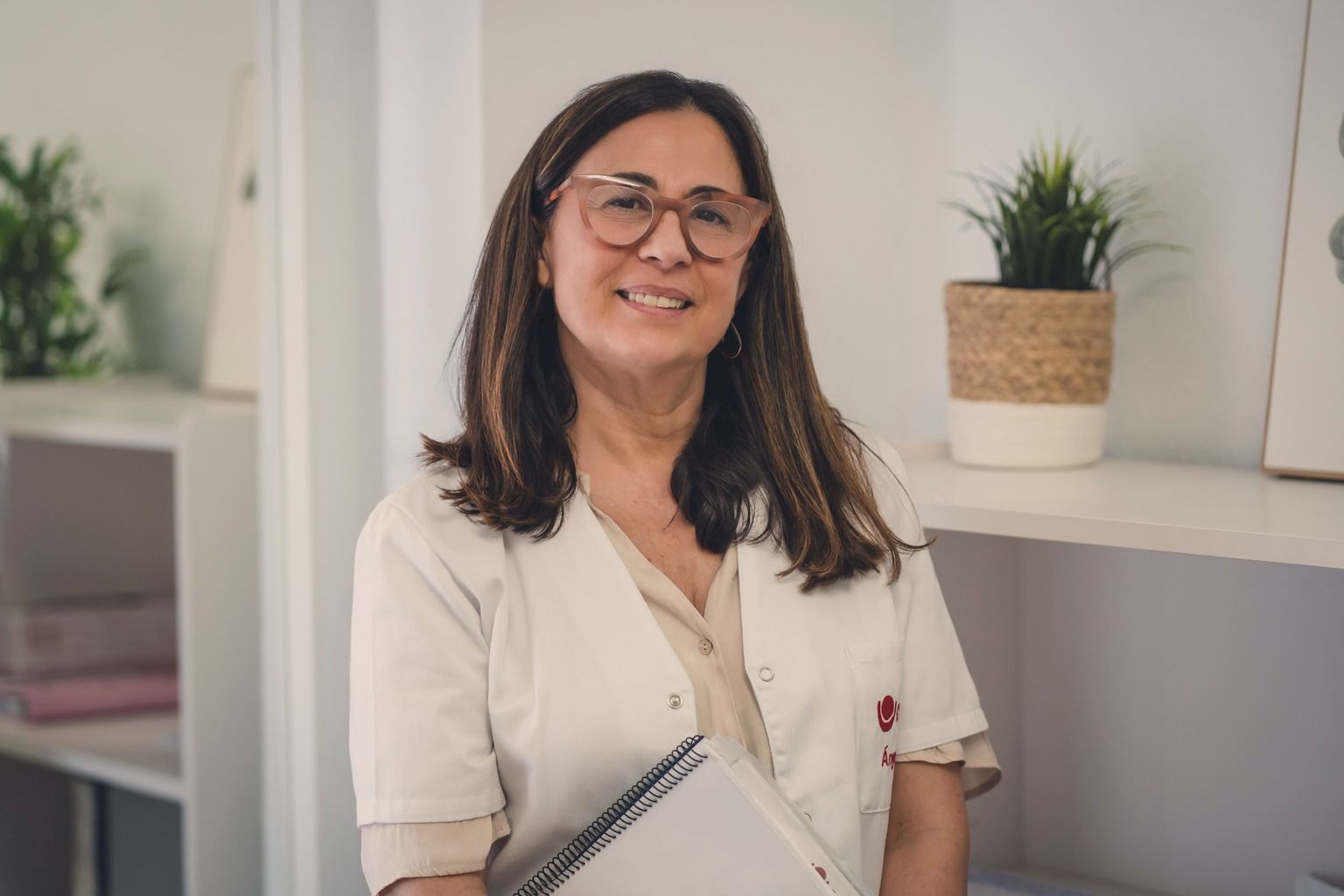 Una mujer con bata de laboratorio y gafas sonríe y sostiene un cuaderno.