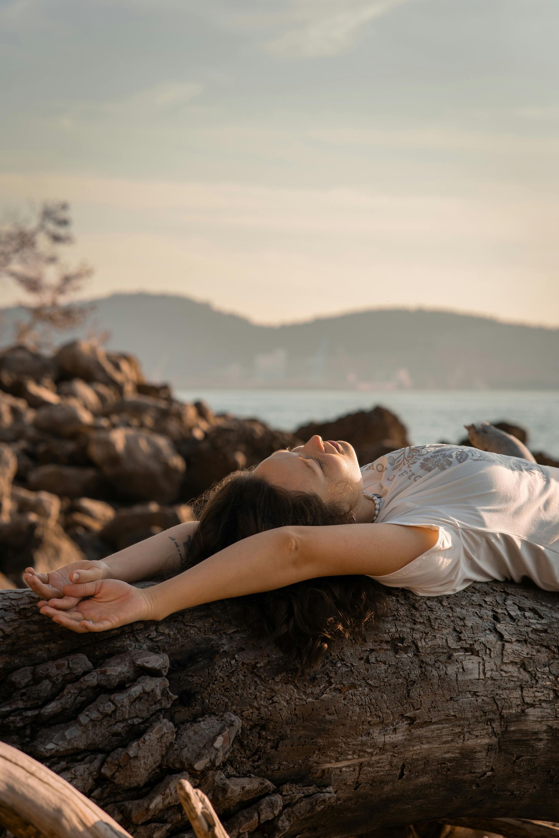 Mujer tumbada sobre un tronco junto al agua al atardecer, con los brazos extendidos sobre la cabeza y las montañas de fondo.