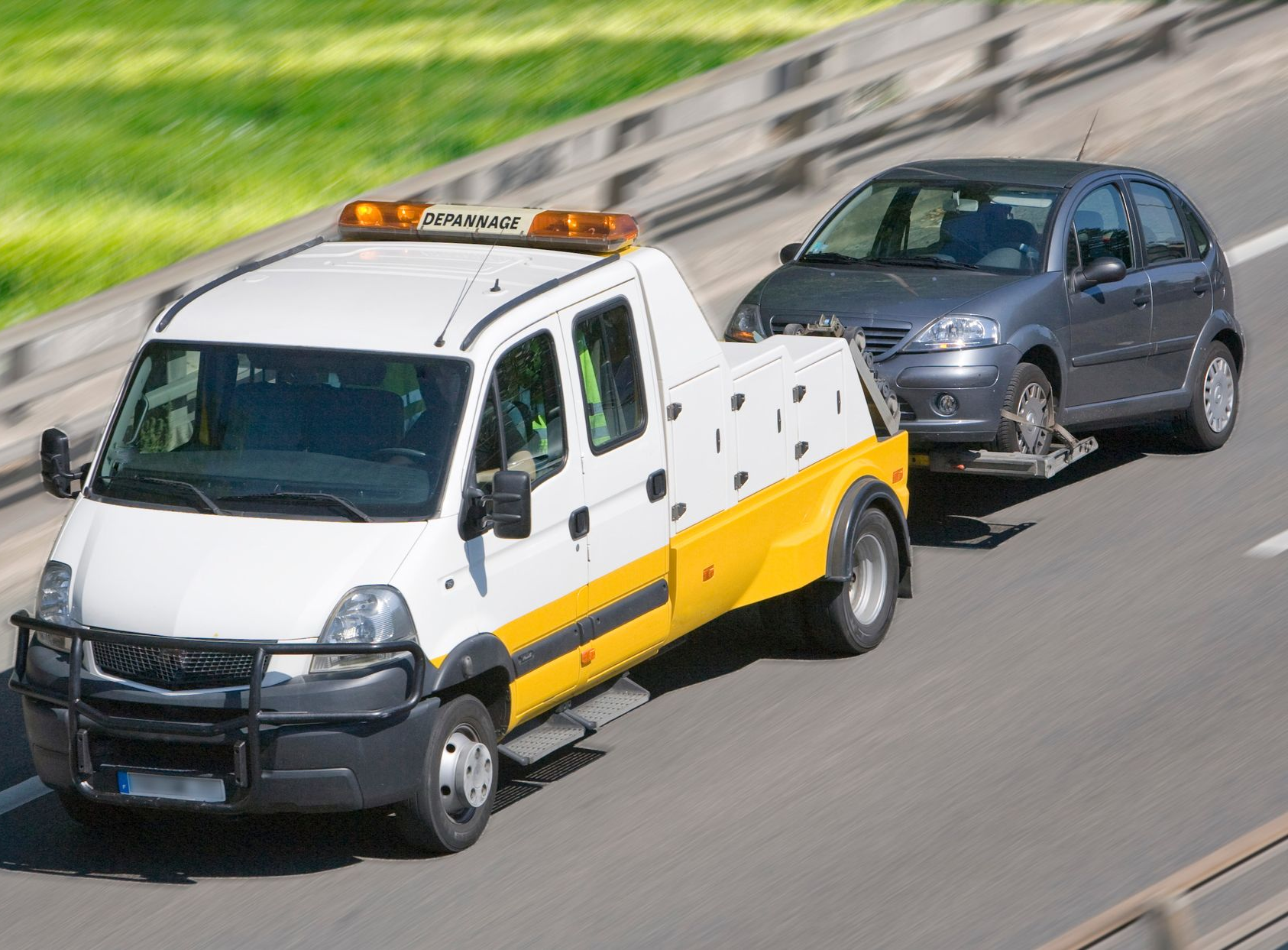 Une dépanneuse remorque une voiture grise sur la route. Le camion est blanc et jaune.