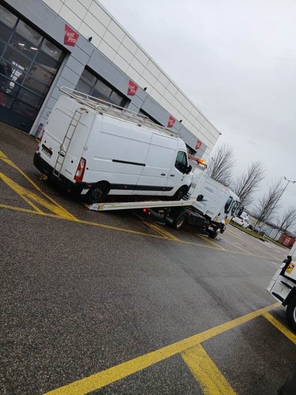 Une dépanneuse remorque une voiture grise ; la dépanneuse est blanche avec des touches de jaune.