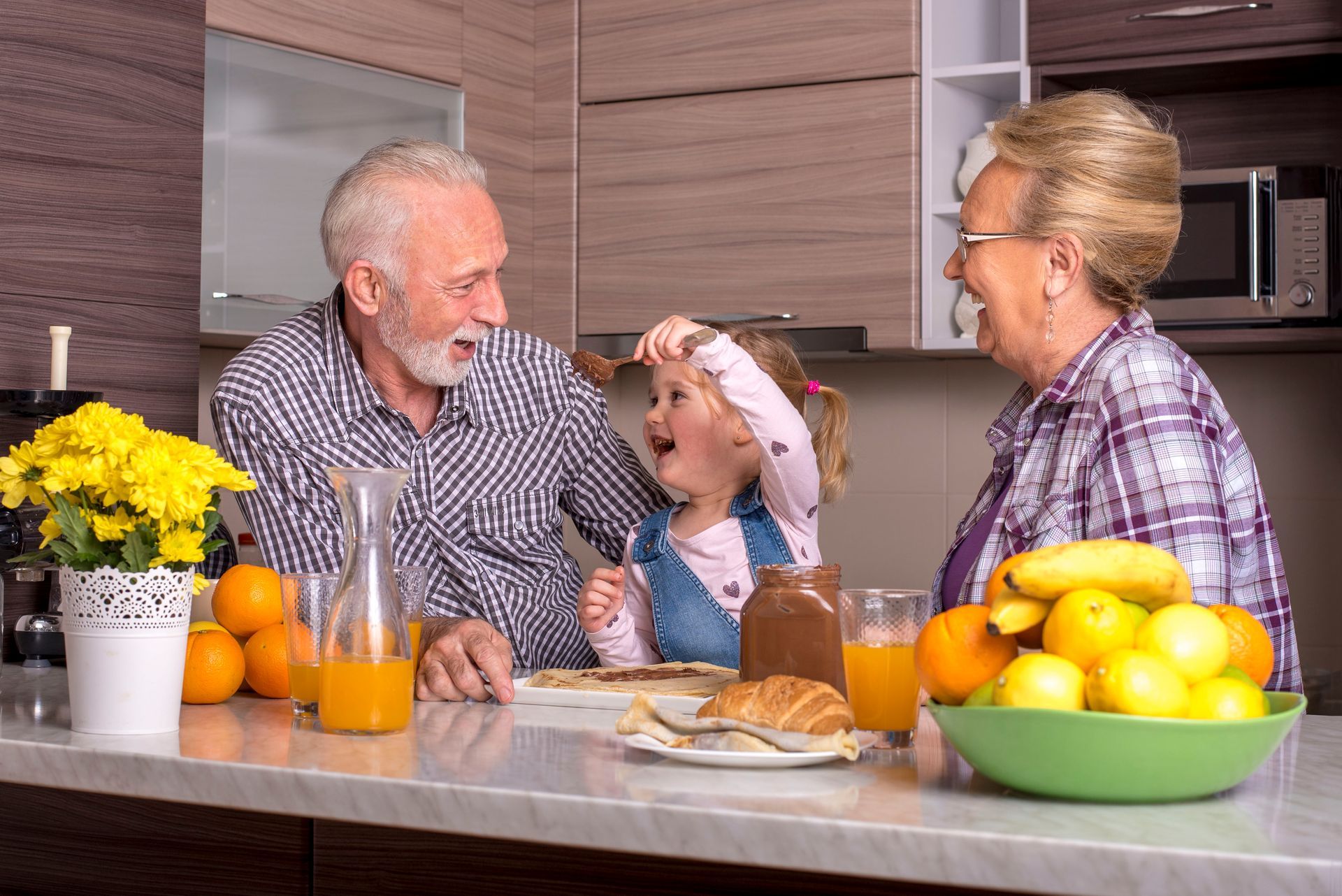 Des grands-parents et un enfant rient ensemble au comptoir de la cuisine, un plat à la main.