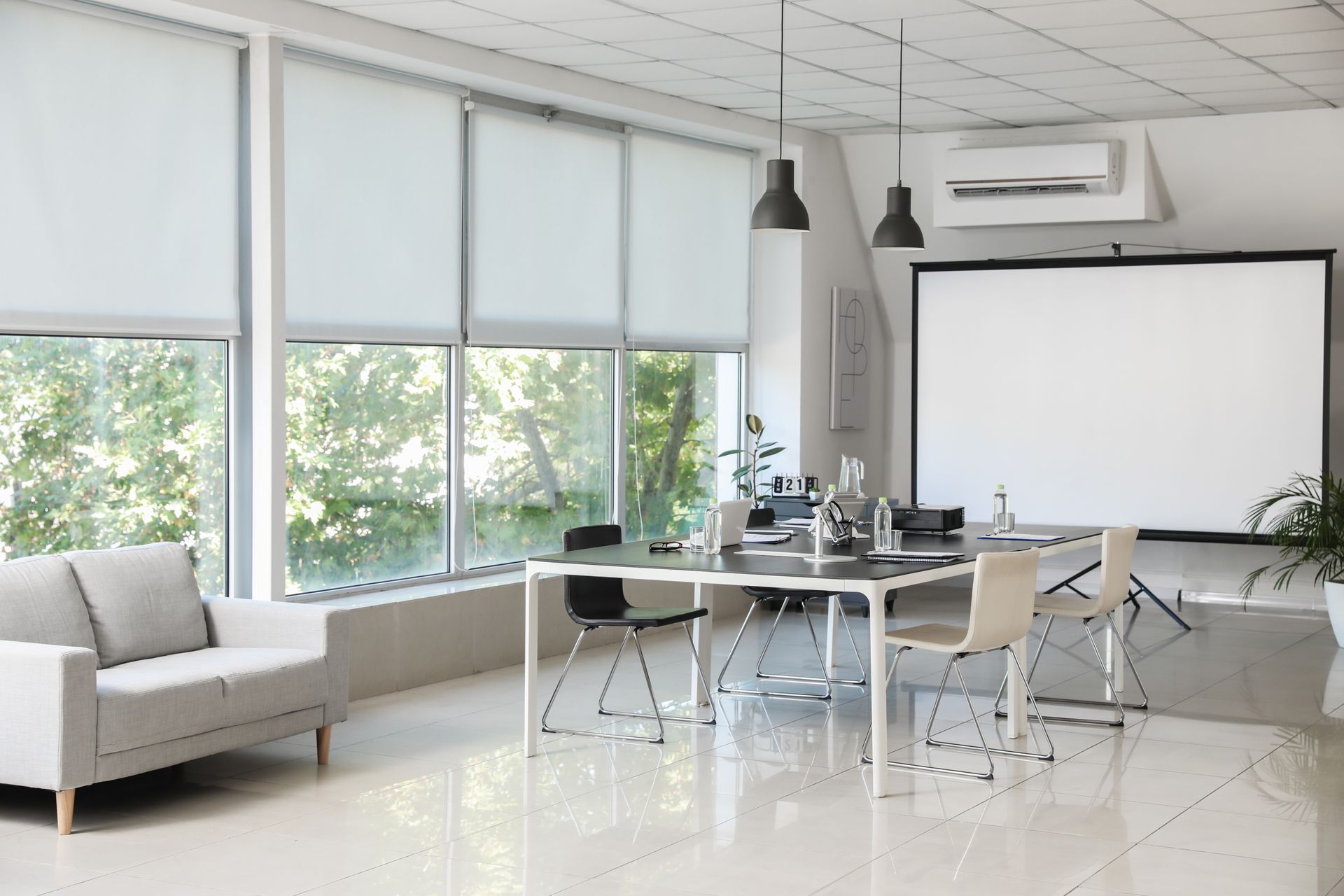 Salle de conférence moderne avec table en verre, chaises blanches, grandes fenêtres et écran de projection.