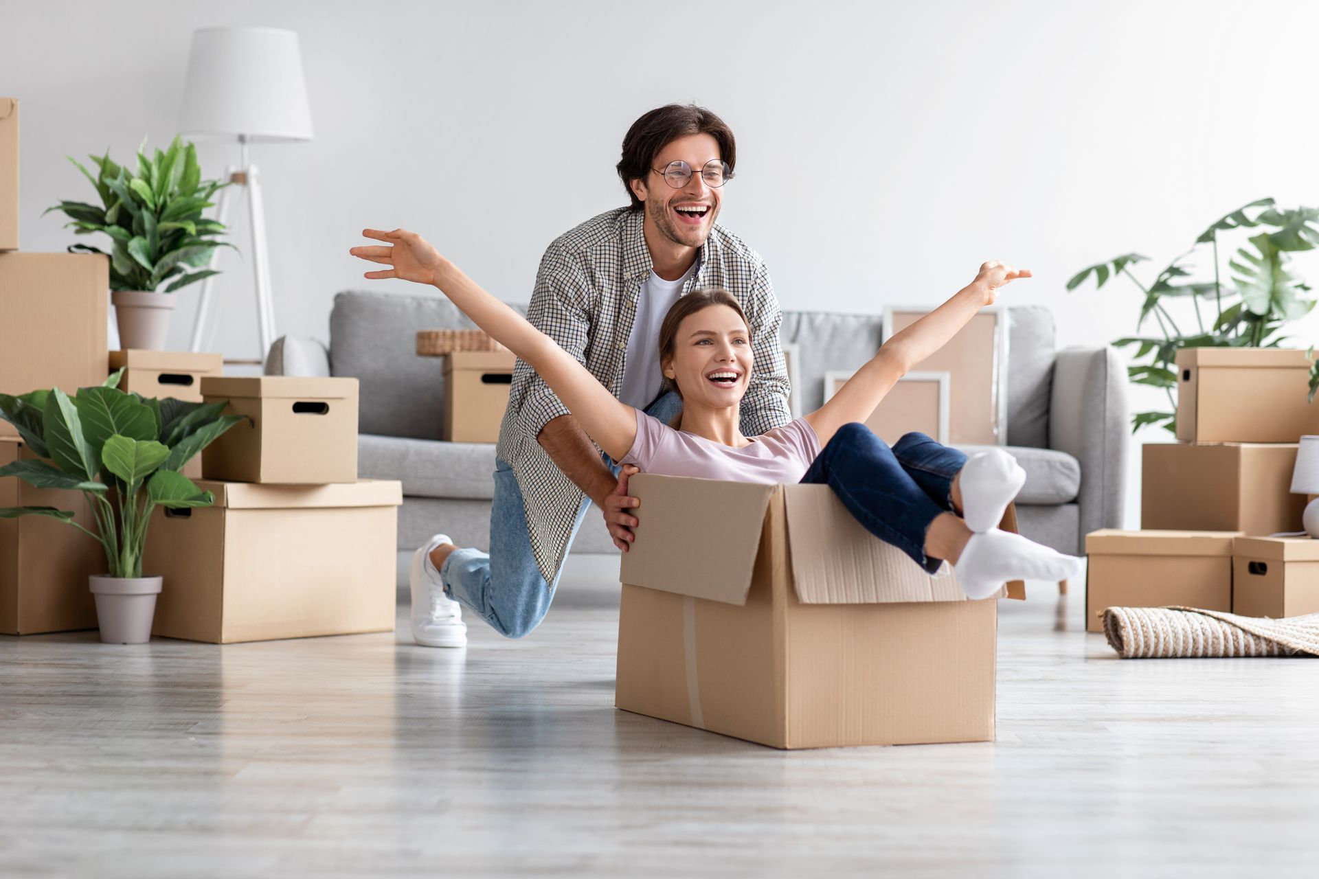 Un homme pousse une femme dans un carton, tous deux rient dans une pièce remplie de cartons de déménagement.