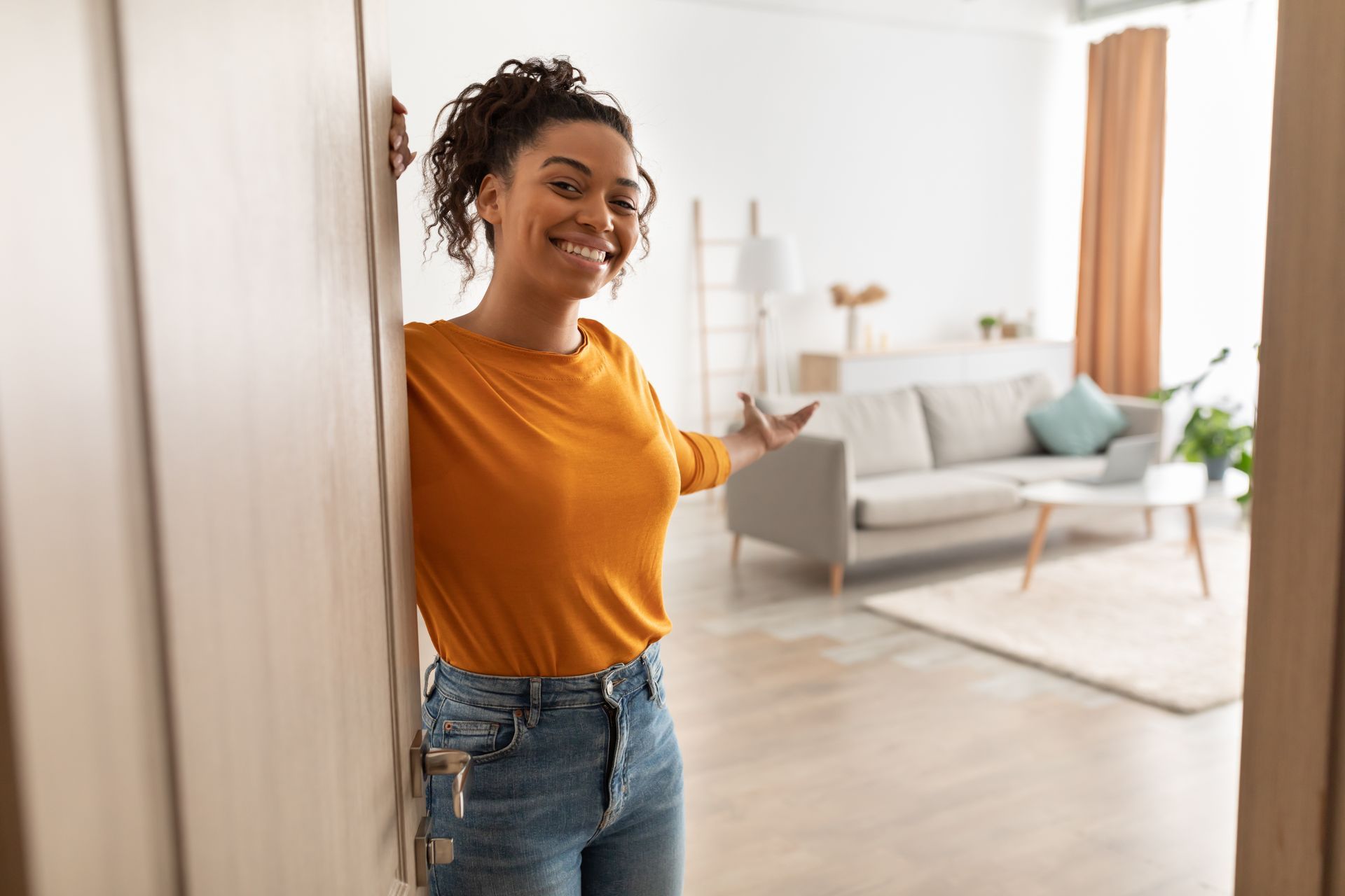 Une femme en chemise orange et jean sourit et désigne son salon.