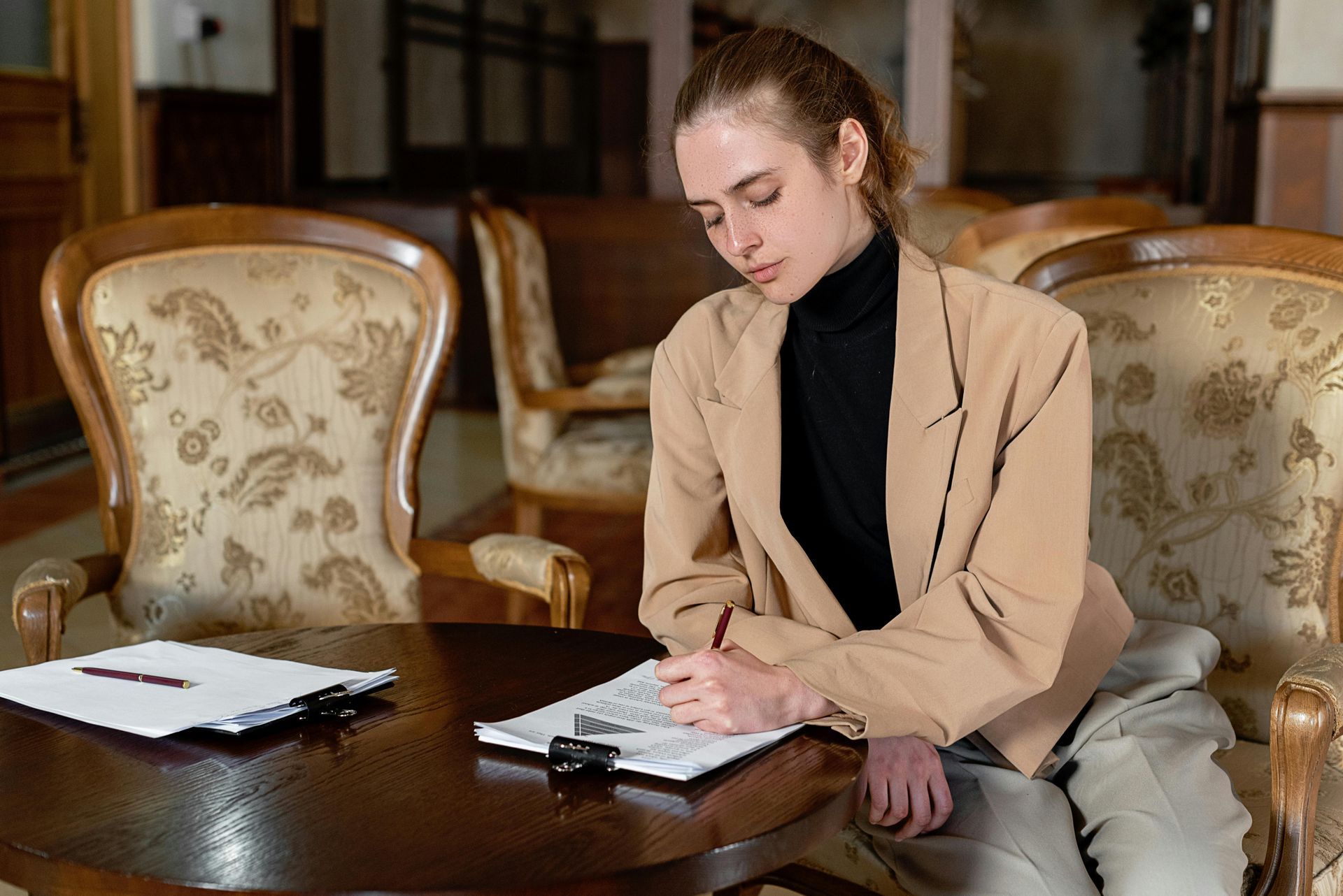 Une femme écrivant à une table dans une pièce, vêtue d'un blazer.