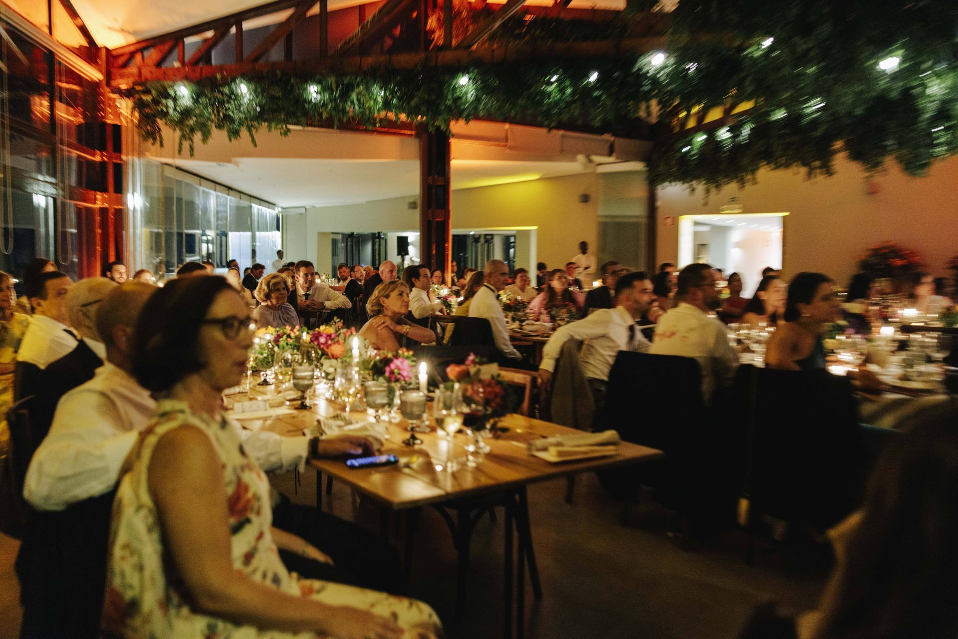 Mão usando relógio e anéis de ouro, segurando uma carteira preta, apoiada sobre a roupa de seda cinza de uma pessoa.