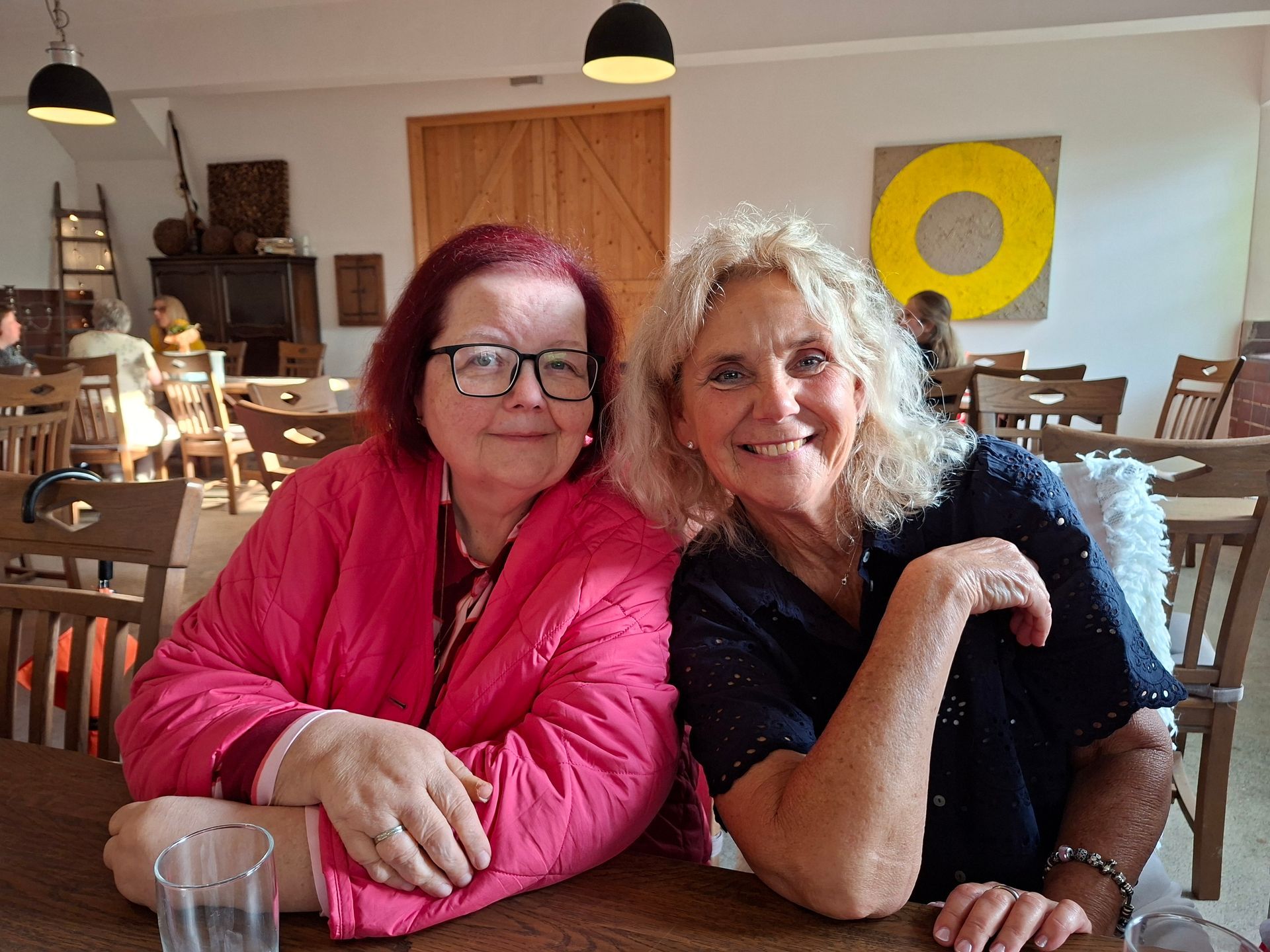 Zwei lächelnde Personen sitzen an einem Café-Tisch, eine trägt eine rosa Jacke, die andere ein dunkelblaues Oberteil mit blauem Schal.