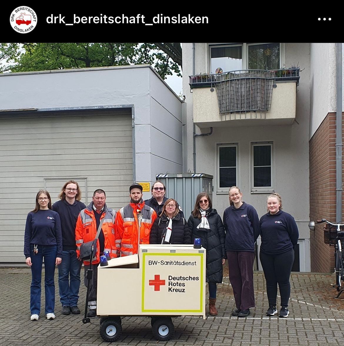Eine Gruppe von Menschen steht um einen Wagen mit einem roten Kreuz darauf