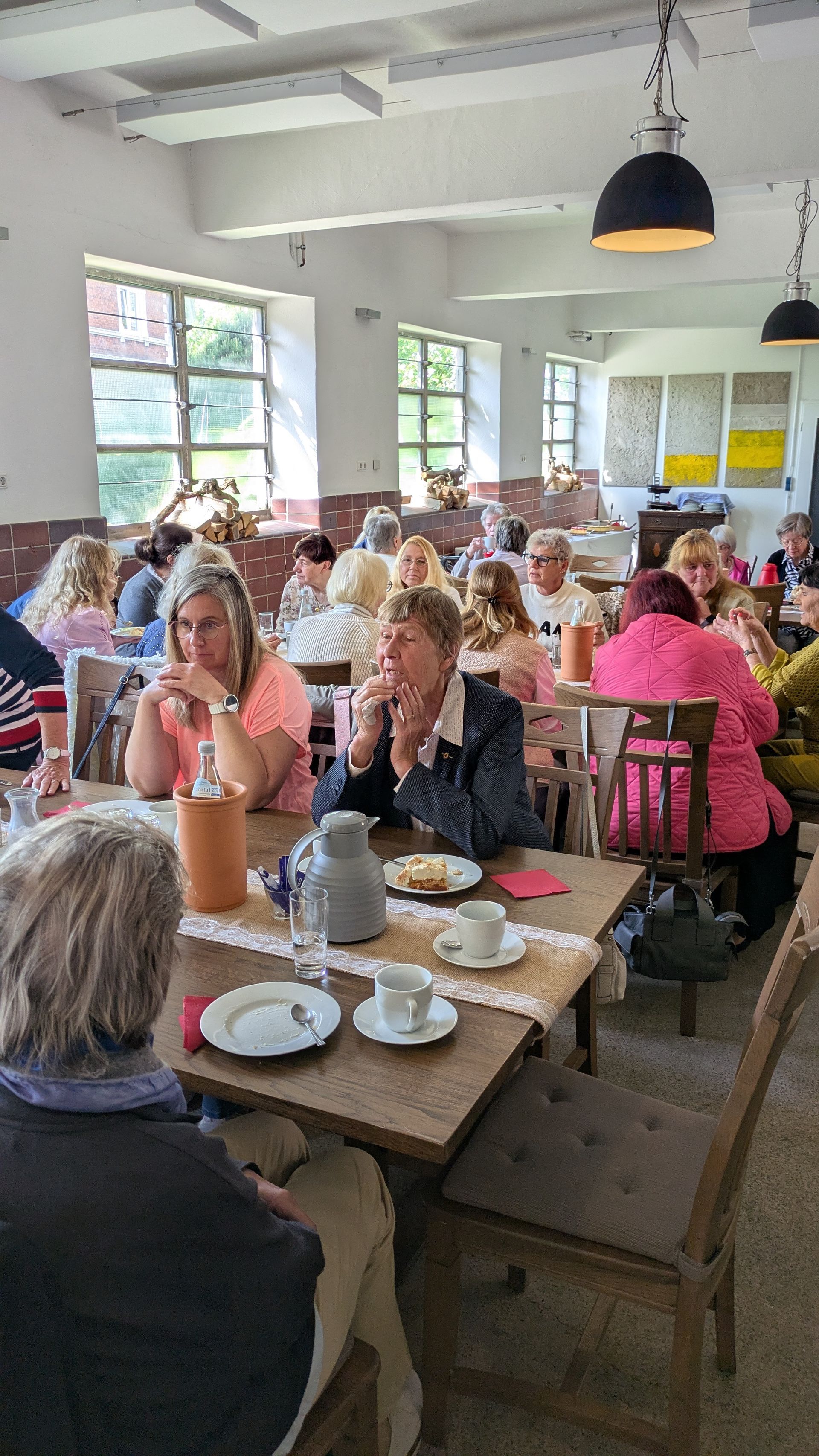 Ein gut besuchtes Café, in dem die Gäste an langen Tischen sitzen, essen und sich unter den Lichterketten unterhalten.