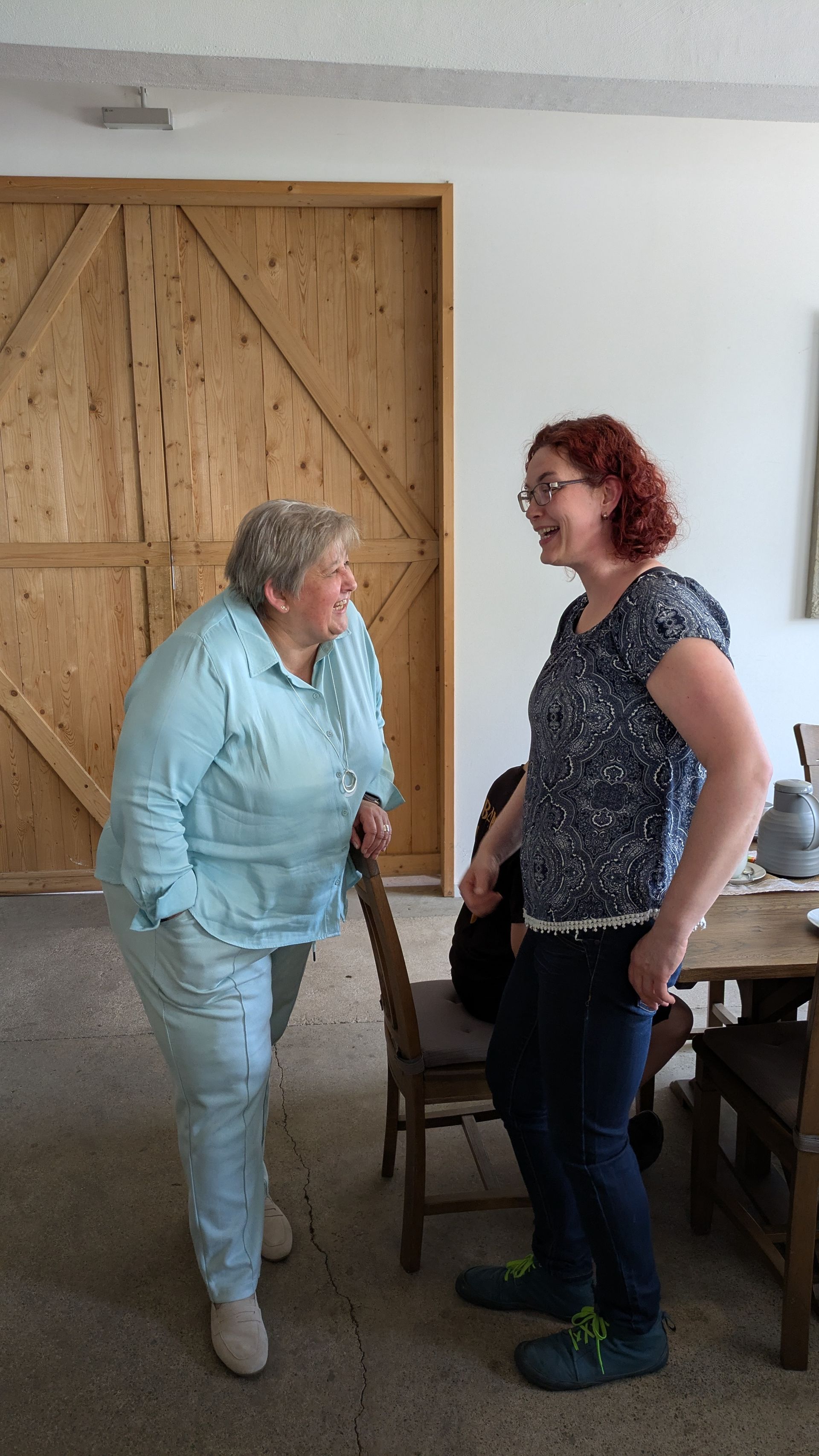 Zwei Personen stehen drinnen neben einer Holztür und einem Esstisch und unterhalten sich.
