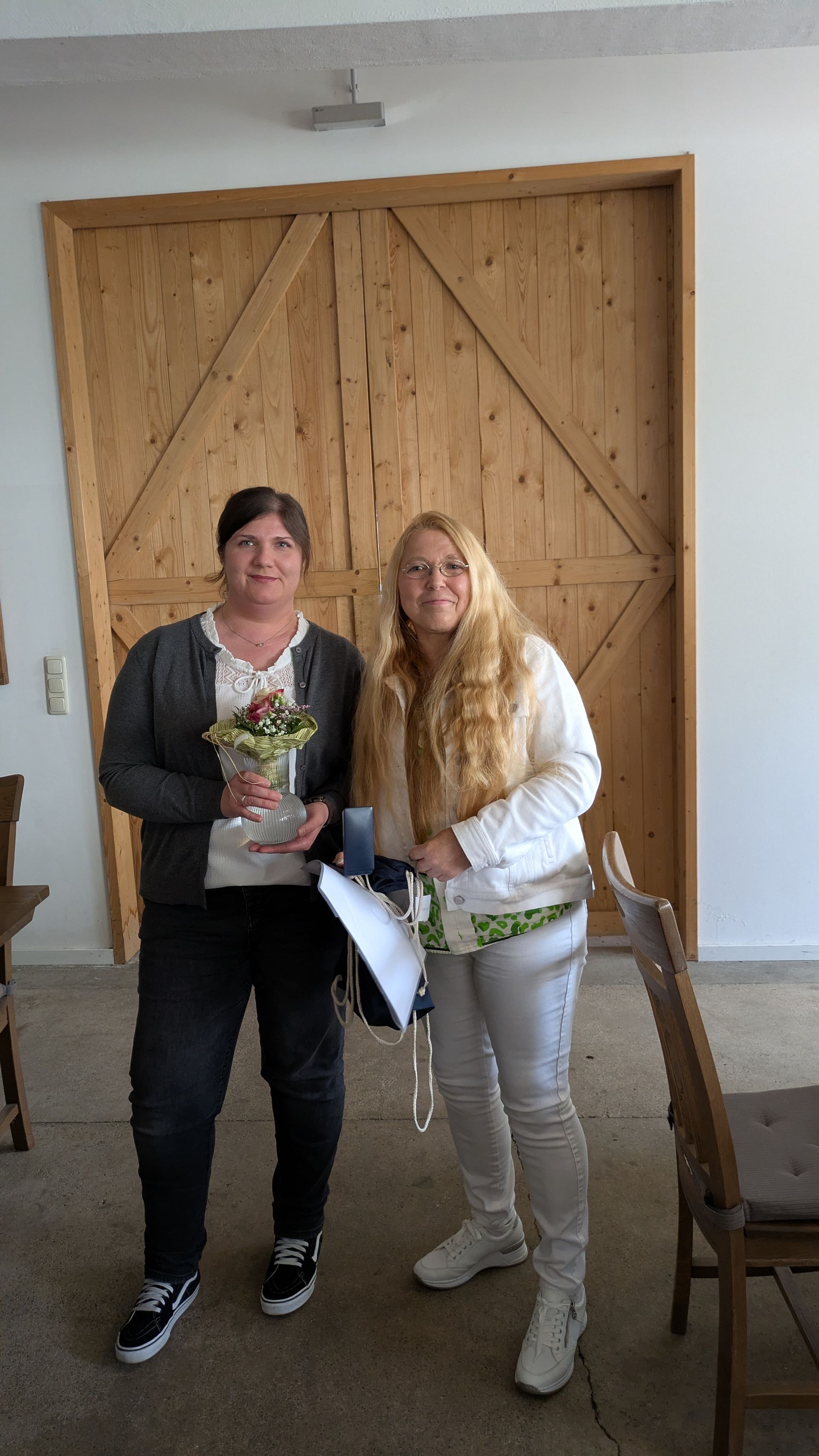 Zwei Personen posieren drinnen vor hölzernen Scheunentoren und halten einen Blumenstrauß und eine Geschenktüte.