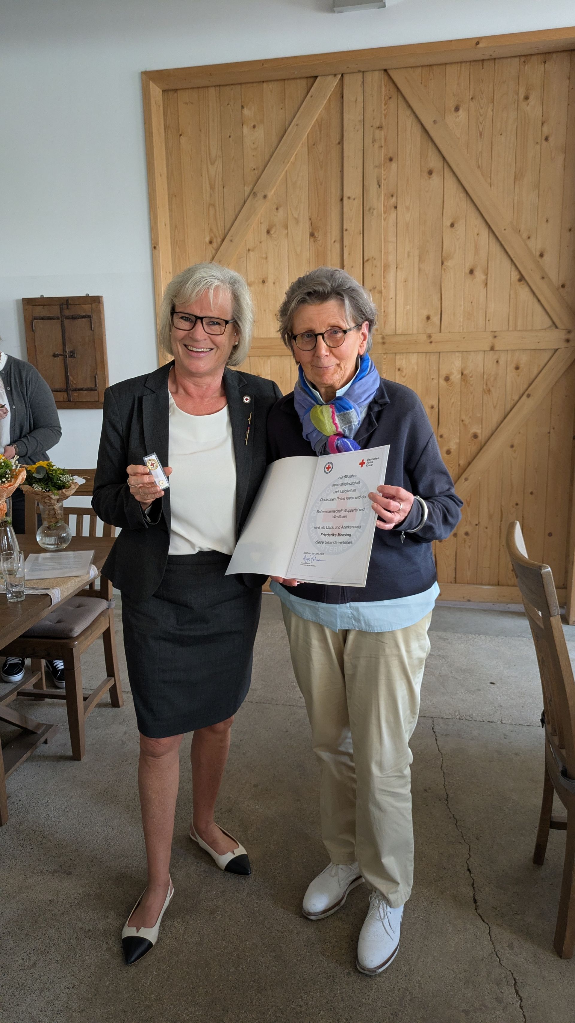 Zwei Frauen halten eine Urkunde und ein Glas und stehen vor einer großen hölzernen Scheunentür.