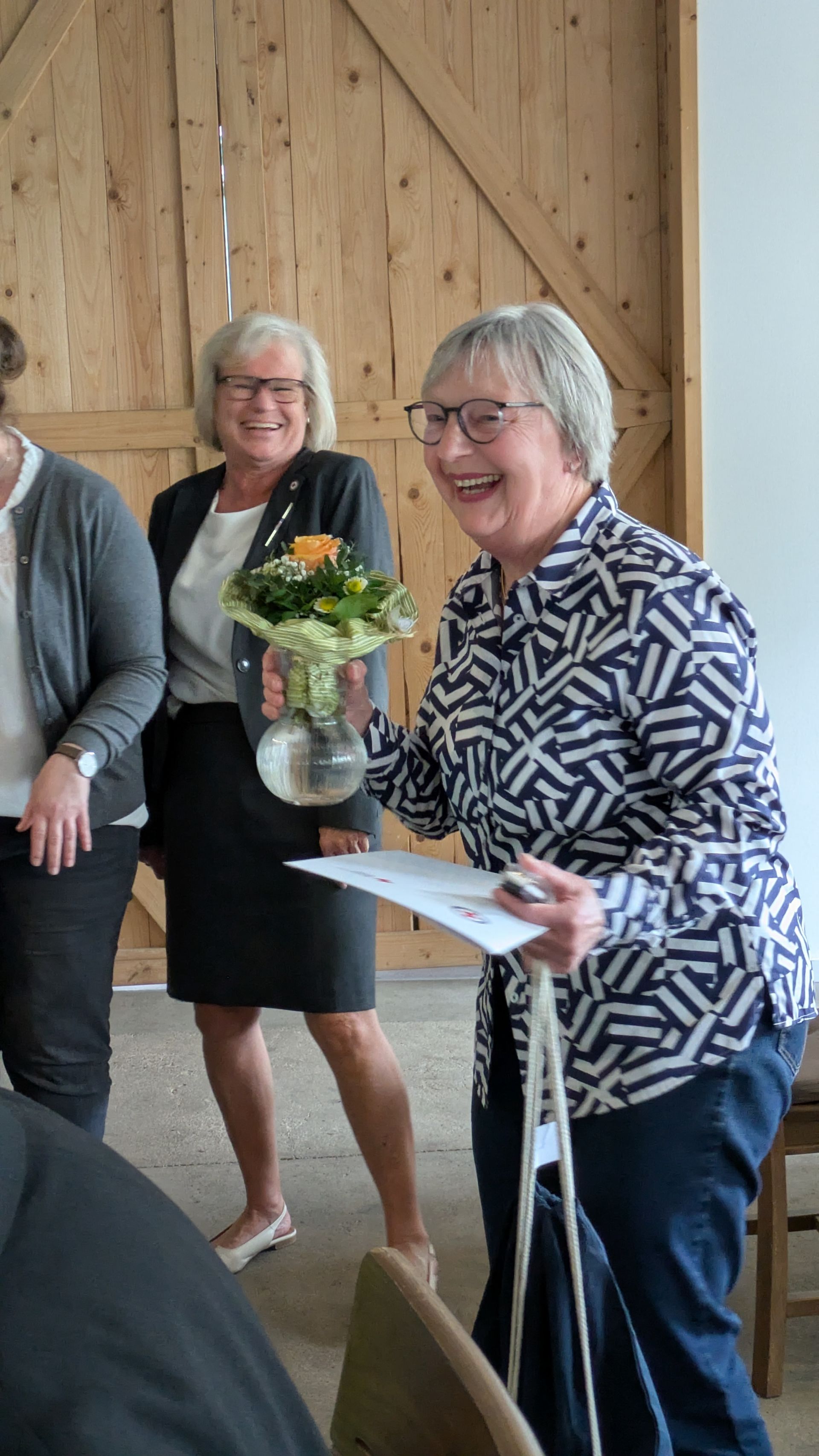 Eine lächelnde Frau in einer gemusterten Bluse hält bei einem Treffen in einem geschlossenen Raum Papiere in der Hand; hinter ihr stehen zwei Frauen und ein Blumenarrangement.